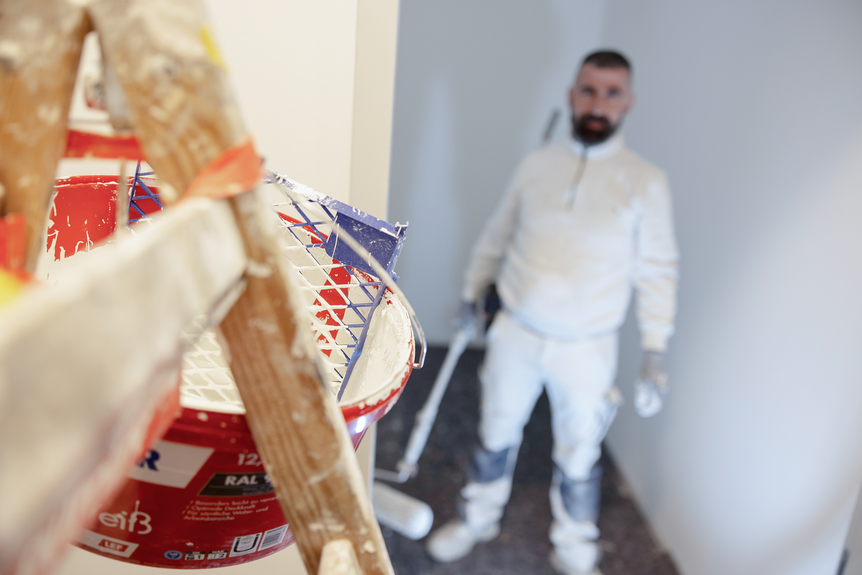 Wichels Malereibetrieb Karriere Maler arbeitet in Innenraum mit Farbrolle und Farbkübel im Vordergrund während Renovierungsarbeiten.